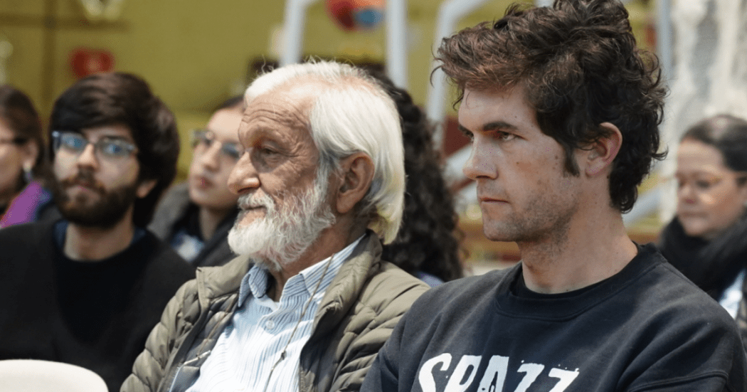 Imagen de varios hombres de diferentes edades sentados prestando atención hacía un escenario.