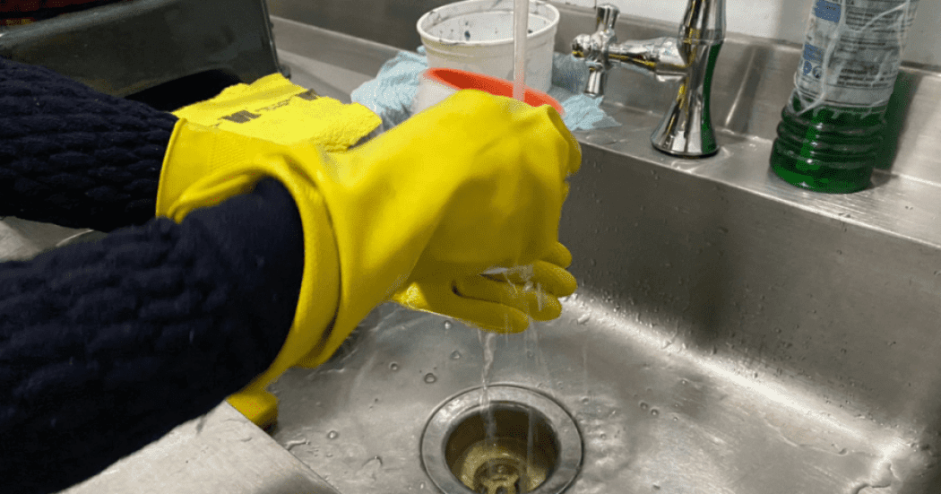 Imagen de unas manos que usan unos guantes amarillos lavando en un lavaplatos gris un vaso de cristal