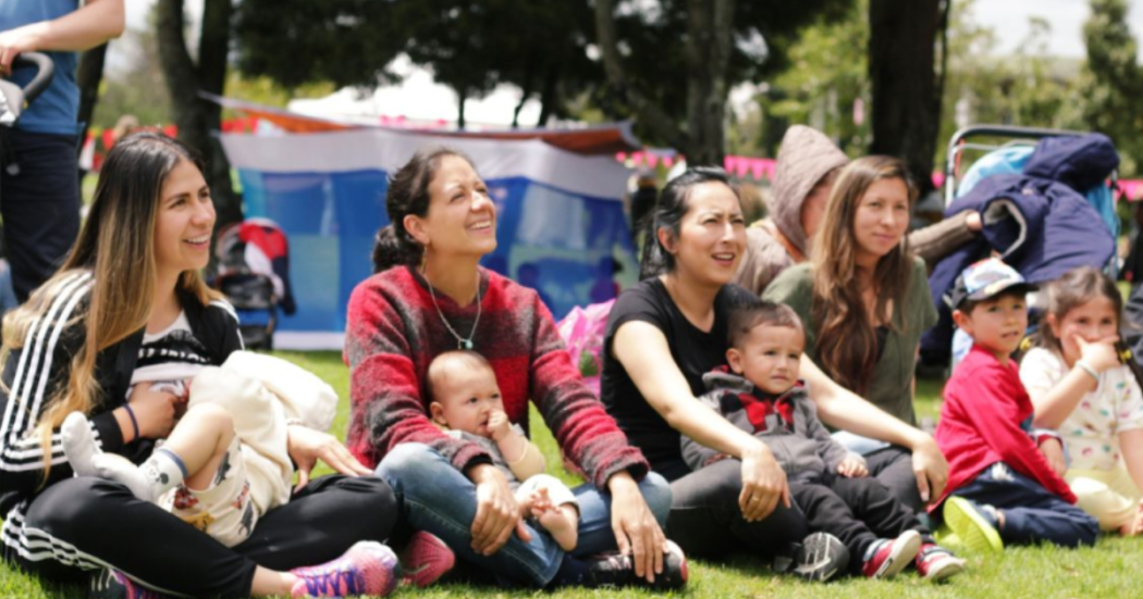 Imagen de mamás con niños en sus piernas sentadas en el pasto en un parque prestando atención a alguien, de fondo se ve la naturaleza del parque