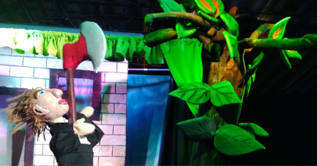 Imagen de un escenario en el que se encuentran un títere de un hombre vestido de chaqueta negra con una hacha en la mano frente a un gran árbol verde.