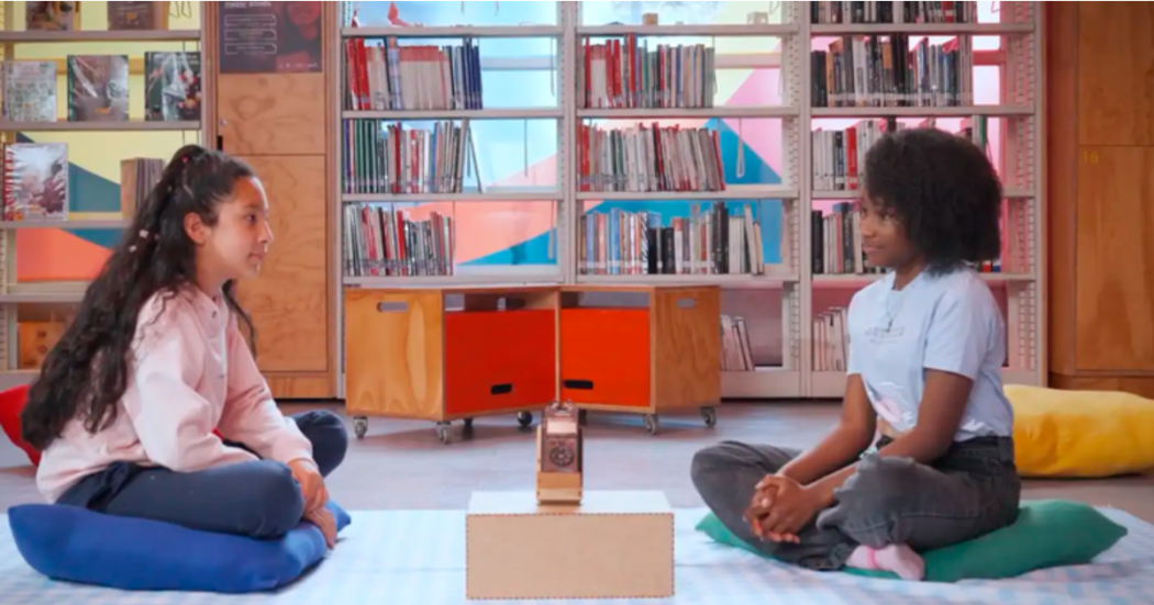 Imagen de dos niñas adolescentes sentadas en el suelo hablando al fondo se ve una biblioteca moderna y muchos libros