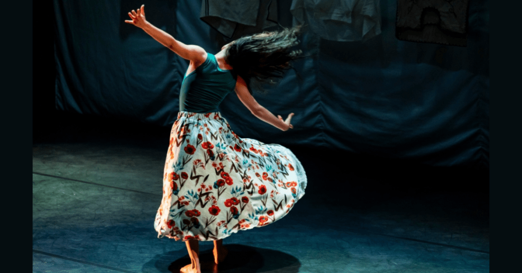 Imagen de una mujer danzando en un escenario cuyo fondo es de color gris oscuro.