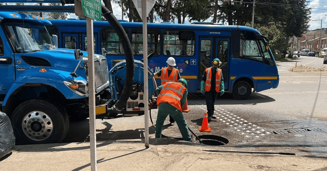 Imagen de tres técnicos de la Empresa de Acueducto realizando mantenimiento en un tubo del acueducto de una calle de la ciudad, al fondo se ven un carrotanque y varios buses del SITP.
