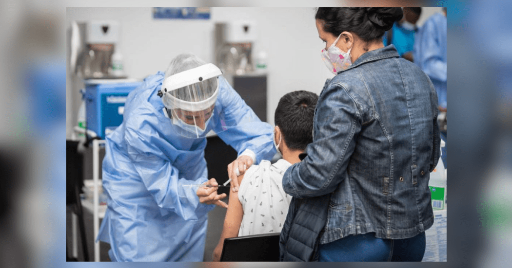 Imagen de una enfermera que vacuna a un niño sentado que es apoyado por una mujer que se encuentra de pie.