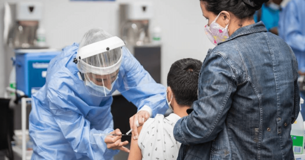 Imagen de enfermera que coloca vacuna a un niño que está sentado en una silla mientras una mujer lo sostiene de su mano..