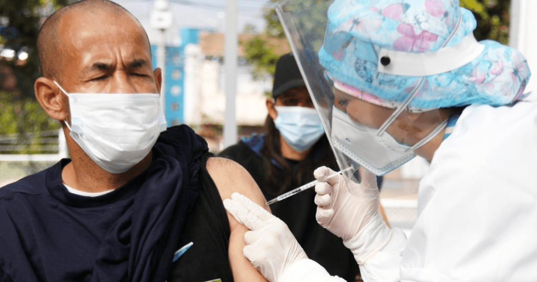Imagen de una mujer con vestido se seguridad aplicando una vacuna a un hombre que está sentada en una silla