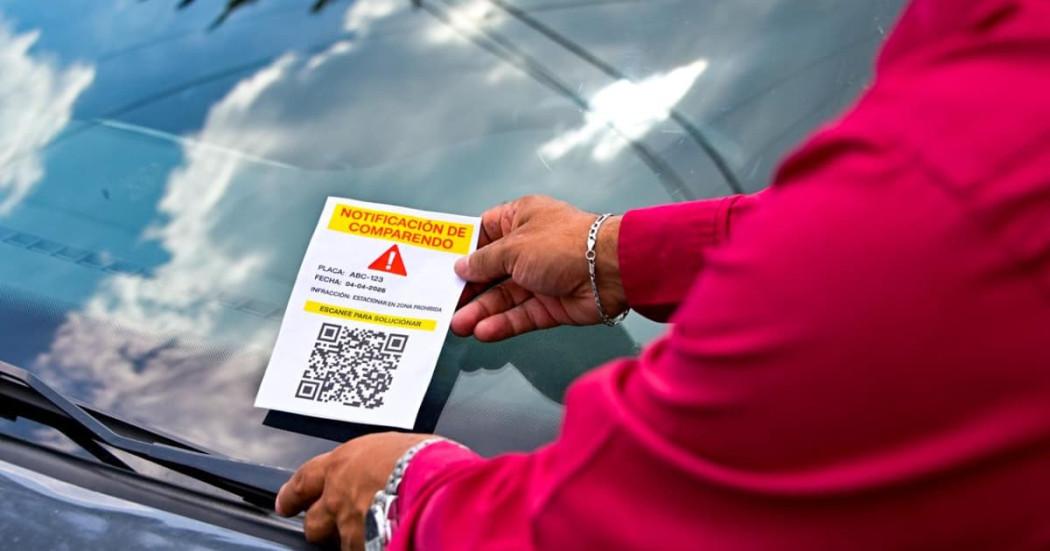 Persona poniendo un volante en el parabrisas de un carro. En el volante se lee: "Notificación de comparendo", con un código QR que invita a escanearse.