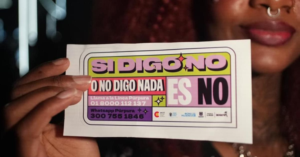 Mano de mujer sosteniendo un volante que dice: "Si digo no o no digo nada, es no. Llama a la Línea Púrpura".