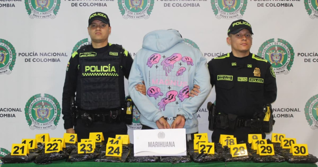 El hombre de la pareja, detenido en medio de dos policías, con el cargamento de marihuana sobre una mesa.