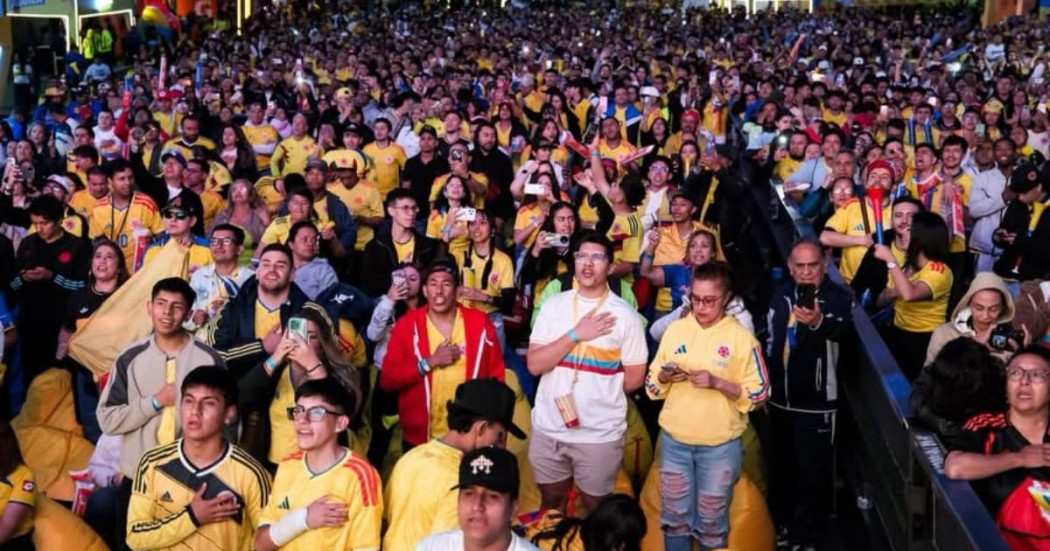 Multitud numerosa reunida en fanzone del Parque El Tunal observa partido de la Selección Colombia en pantallas gigantes; asistentes visten camisetas amarillas, sostienen banderas, teléfonos móviles y aplauden de pie en jornada nocturna.