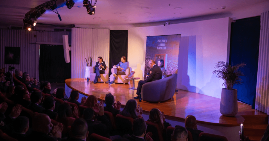 Panel de conversación en auditorio con cuatro personas sentadas en el escenario frente a público asistente, iluminación azul y morada, plantas decorativas y banner al fondo.