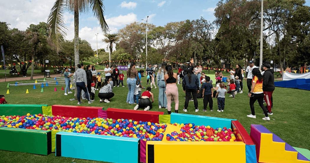 Personas adultas y menores participan en actividades recreativas sobre una zona verde de parque; en primer plano hay piscinas secas con pelotas de colores y al fondo se observan conos, árboles y grupos reunidos al aire libre.