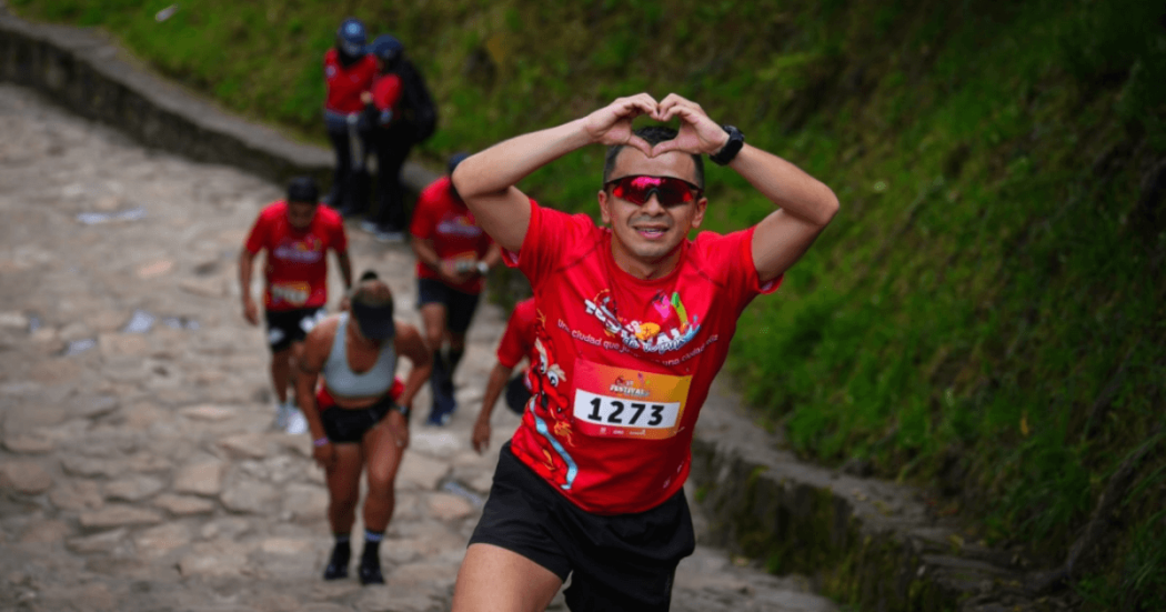 Participantes ascienden por el sendero de Monserrate, en primer plano un corredor con el número 1273 realiza un gesto de corazón con las manos mientras continúa el recorrido.
