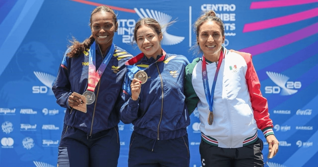 Gabriela Rueda exhibe su medalla de oro durante ceremonia de premiación de los Juegos Centroamericanos y del Caribe Santo Domingo 2026, frente al panel oficial del evento deportivo.
