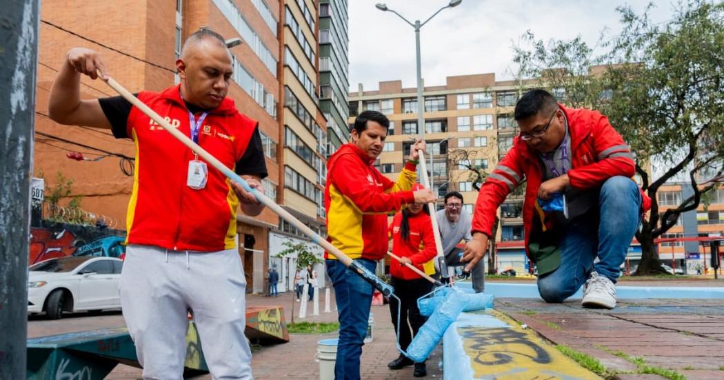 Grupo de trabajadores realiza labores de mantenimiento urbano pintando bordillos y superficies peatonales en espacio público, utilizando rodillos y pintura azul, con edificios residenciales y zona vial visibles al fondo.