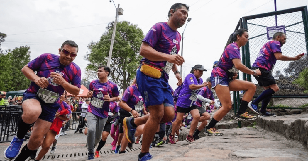 Imagen de referencia sobre participantes de la carrera Sube Monserrate ascienden al inicio del recorrido, vistiendo camisetas moradas del evento y números de competencia visibles en el pecho.