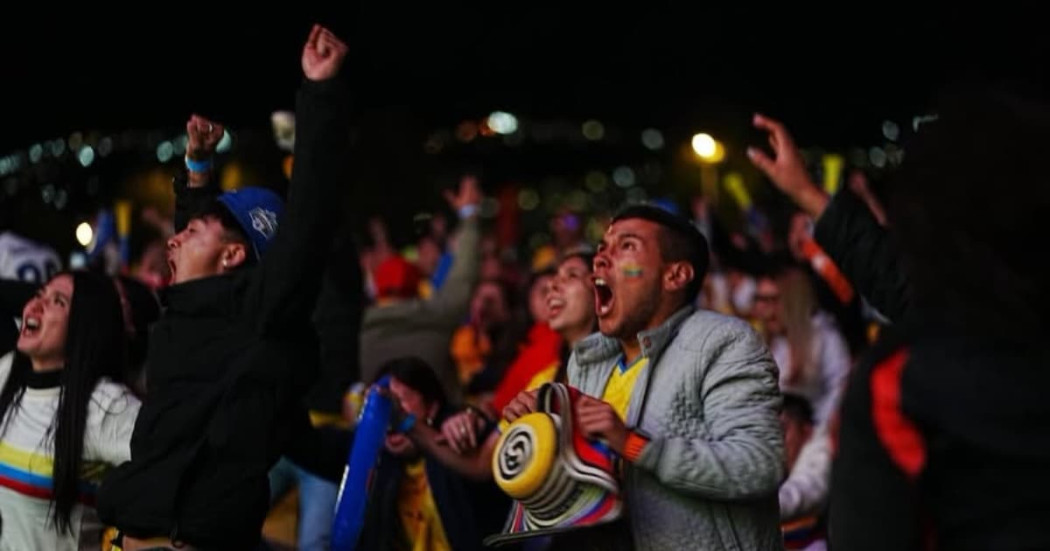 Multitud reunida en fanzone nocturno de Bogotá observa partido en pantallas gigantes, con asistentes levantando brazos, vistiendo prendas de la Selección Colombia