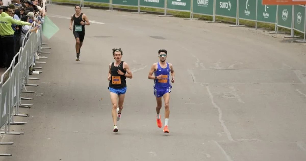 Grupo de atletas corre en la Media Maratón de Bogotá por una vía amplia durante competencia atlética urbana, con vallas metálicas a ambos lados y público observando desde el costado izquierdo