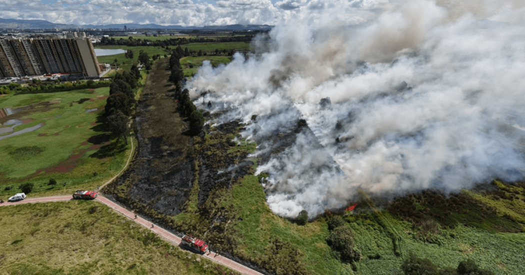 Vista aérea del humedal Jaboque durante un incendio de cobertura vegetal, con extensas columnas de humo, áreas afectadas por el fuego, vehículos de emergencia y edificaciones cercanas.