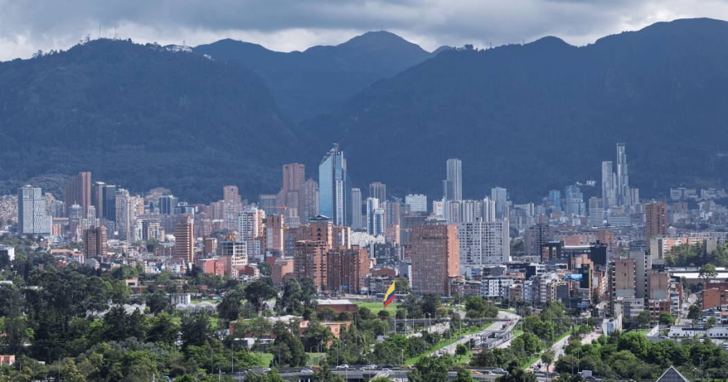 Vista panorámica de Bogotá con edificios residenciales y empresariales, zonas verdes, la bandera de Colombia en primer plano y los cerros orientales bajo un cielo nublado.