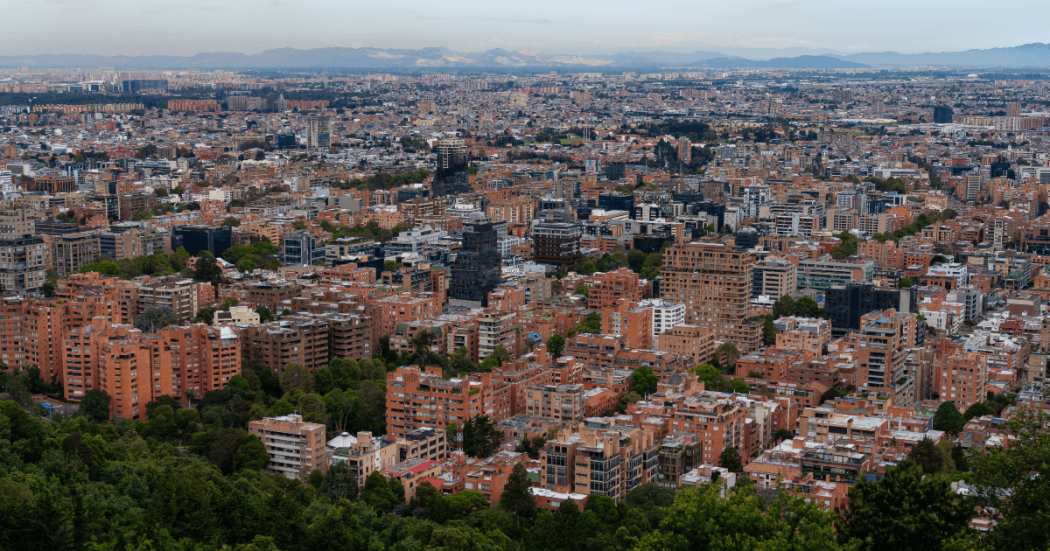 Vista panorámica de Bogotá con alta concentración de edificios residenciales y comerciales, amplias zonas arboladas en primer plano y expansión urbana que se extiende hacia el occidente de la ciudad.
