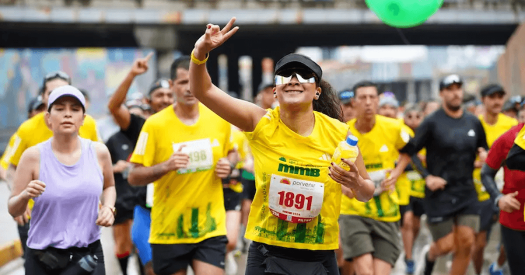 Corredores participan en la Media Maratón de Bogotá mientras avanzan por una avenida de la ciudad; en primer plano una atleta con camiseta amarilla y número 1891 saluda al público durante la competencia.