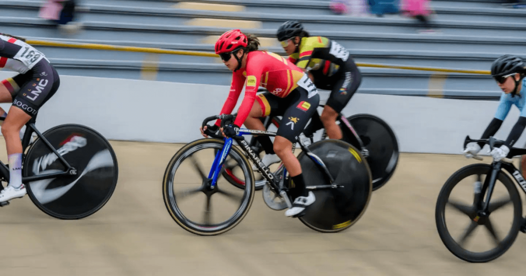 Ciclistas compiten en una prueba de ciclismo de pista sobre un velódromo. En primer plano se observan bicicletas de pista con ruedas lenticulares, cascos y uniformes deportivos de diferentes colores.