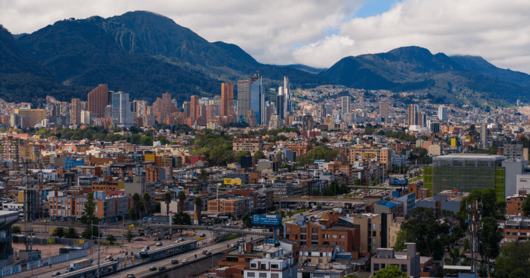 Vista panorámica de Bogotá con edificaciones residenciales y comerciales, vías principales y los cerros orientales al fondo bajo un cielo parcialmente nublado.