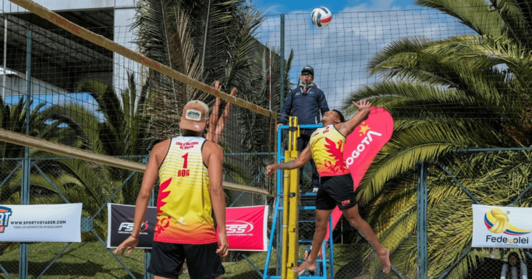 Partido de voleibol playa con dos deportistas junto a la red durante competencia al aire libre, con palmeras y juez en silla de arbitraje.
