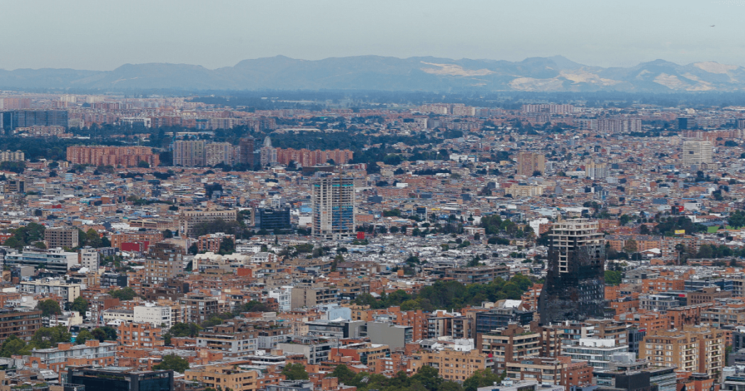 Vista panorámica de Bogotá con gran concentración de edificaciones residenciales y comerciales, vías urbanas y montañas al fondo en un día despejado.