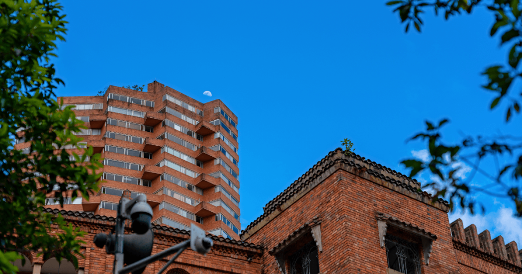 Edificaciones de ladrillo y torre de apartamentos en Bogotá, con vegetación en primer plano y cielo azul al fondo, en una escena urbana residencial.