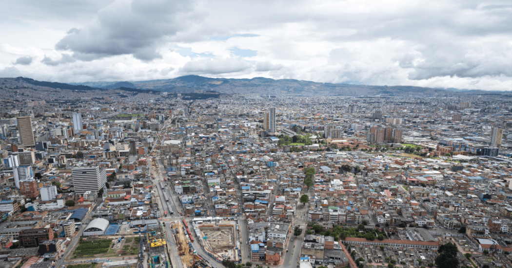 Vista aérea de Bogotá con alta concentración de edificaciones, corredores viales, zonas en construcción y montañas al fondo bajo un cielo nublado.