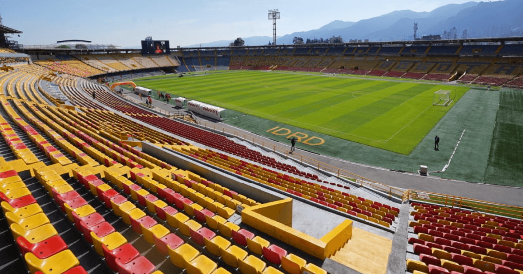 Vista interior del estadio El Campín en Bogotá con graderías amarillas y rojas, cancha de fútbol en óptimas condiciones y montañas al fondo.