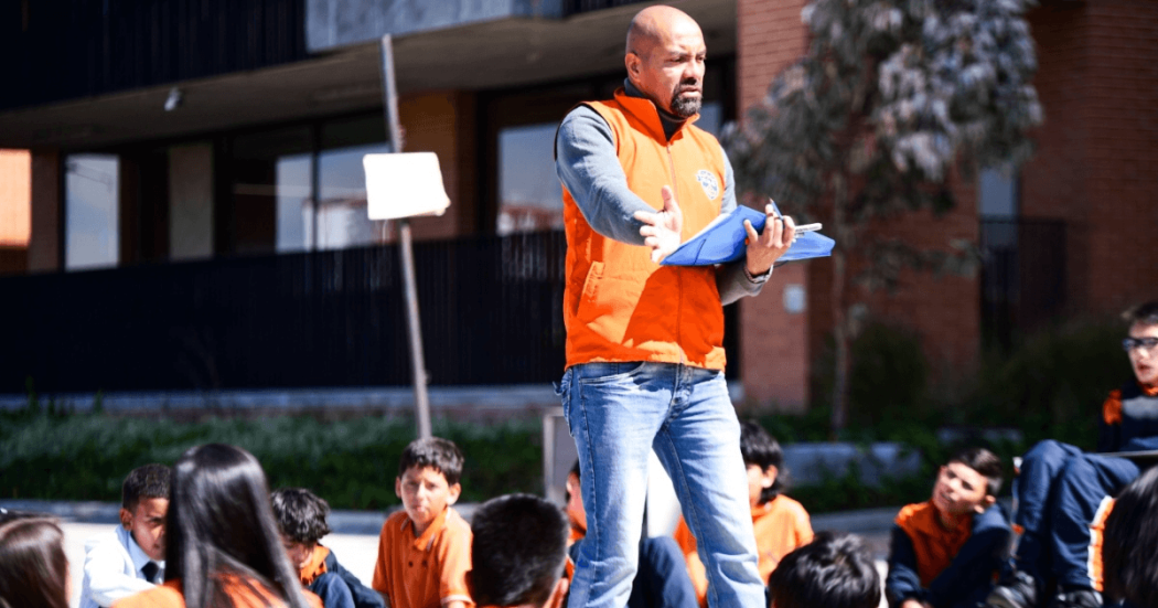 Foto que muestra un hombre con una carpeta en las manos y estudiantes sentados en el piso 