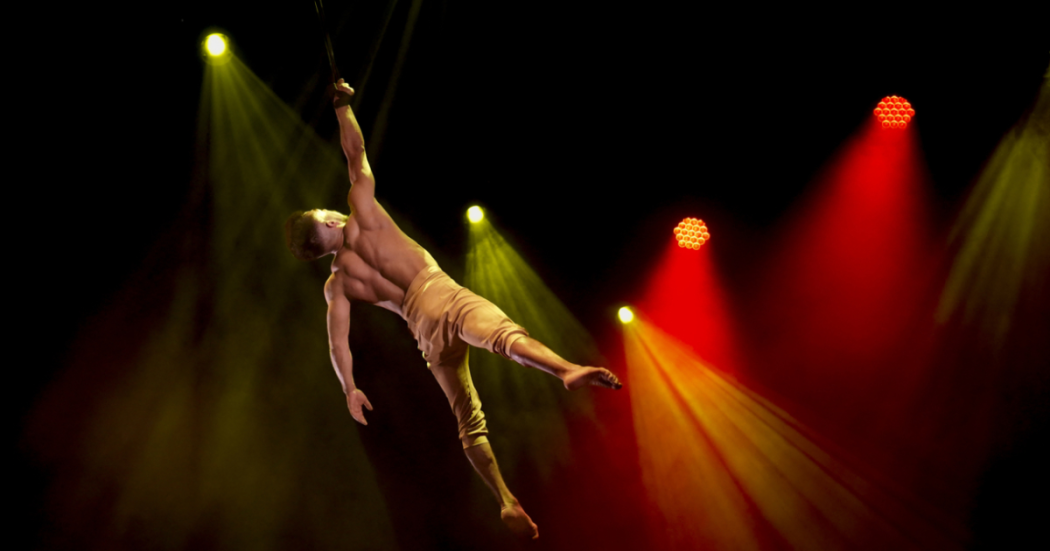 Imagen de un artista de circo en una puesta en escena al aire, de fondo se ven lices blancas y amarillas.