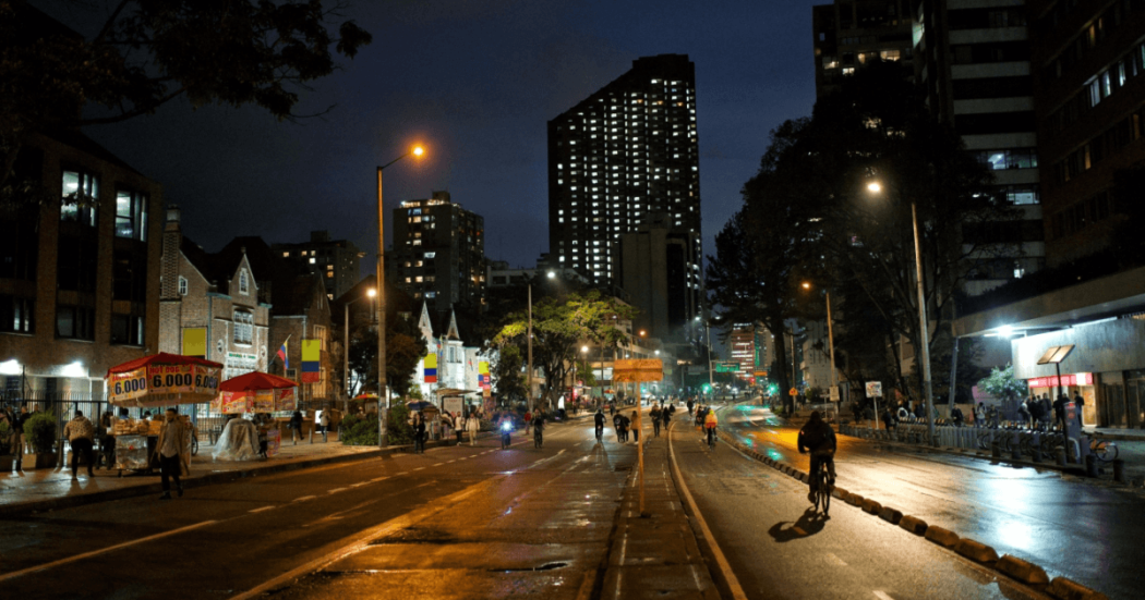 Ciclistas y peatones recorren una avenida iluminada durante la Ciclovía Nocturna de Bogotá, con edificios, comercio, alumbrado público y pavimento húmedo visibles en horario nocturno.