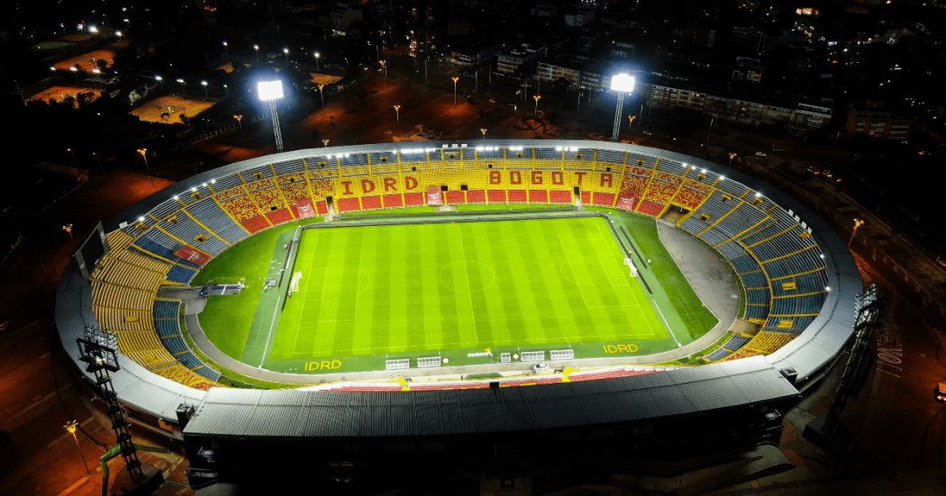 Vista aérea nocturna del estadio El Campín en Bogotá, iluminado por reflectores, con gramilla, graderías multicolores y entorno urbano visible alrededor.