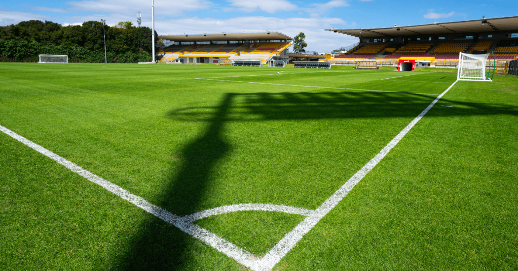 Cancha de fútbol del estadio de Techo con césped renovado, líneas demarcadas, arcos y graderías vacías bajo cielo parcialmente despejado.
