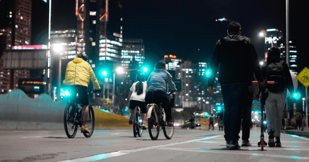 Personas en bicicleta y caminando durante la Ciclovía Nocturna en una vía de Bogotá iluminada por luces urbanas.