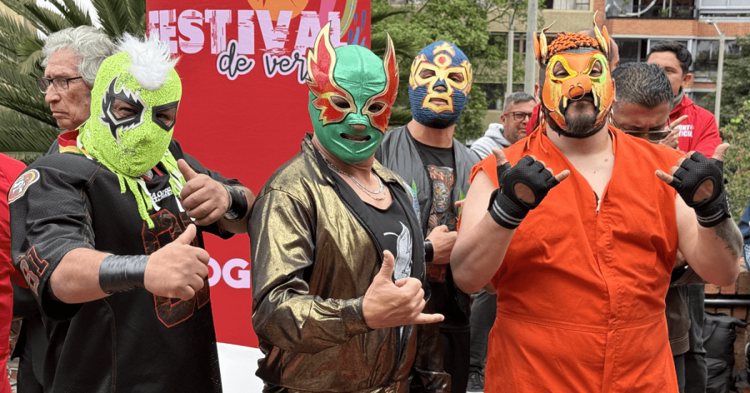 Cuatro luchadores con máscaras coloridas posan frente a un aviso del Festival de Verano durante una actividad de lucha libre en Bogotá.