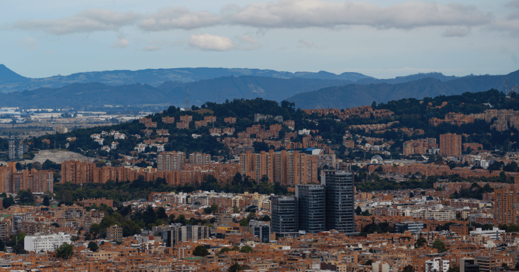 Vista panorámica de Bogotá con edificaciones residenciales y comerciales, zonas arborizadas y cerros al fondo bajo un cielo parcialmente nublado.