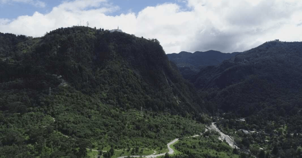 Vista aérea de los Cerros Orientales de Bogotá con laderas cubiertas de bosque, un valle atravesado por una vía y una quebrada.