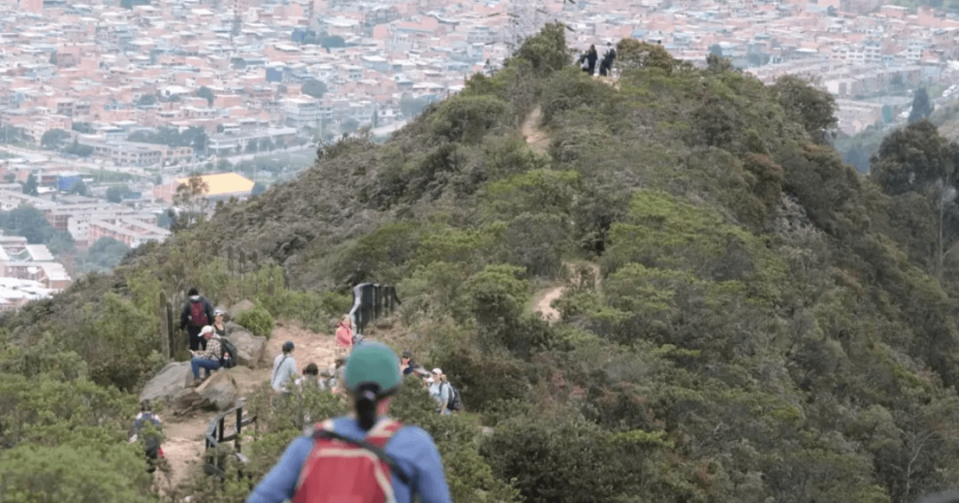 senderos del Parque de Montaña Entrenubes que es recorrido por personas en un día soleado. Al fondo de la imagen se observa una vista panorámica de Bogotá.