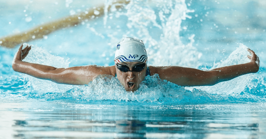 Persona practica natación estilo mariposa en una piscina de Bogotá, con gafas y gorro de natación mientras avanza entre salpicaduras de agua.