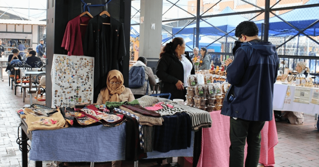 Foto que muestra un grupo de personas en una feria donde se exhiben artesanías y prendas de vestir. 