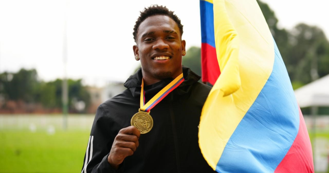 Ronal Longa sosteniendo una medalla dorada colgada al cuello y una bandera de Colombia. Al fondo se observa una pista o campo deportivo con vegetación desenfocada.