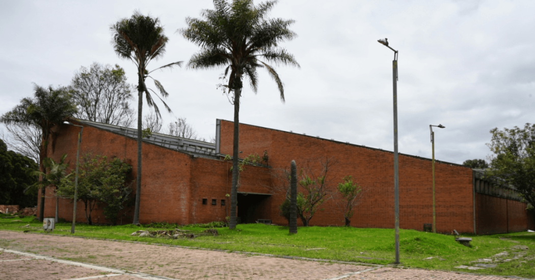 Fachada de ladrillo de una edificación rodeada de zonas verdes, con varias palmas y plantas ornamentales sembradas frente al muro.