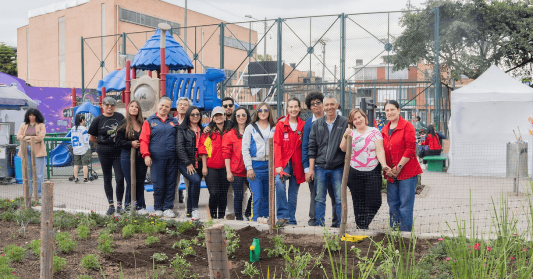 Grupo de personas con chaquetas rojas y ropa casual de pie junto a un jardín recién sembrado, frente a una estructura metálica y edificaciones del sector. En primer plano se observan plantas jóvenes y áreas con tierra.