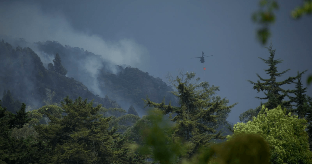 Helicóptero con dispositivo para descarga de agua sobre una zona montañosa con presencia de humo, mientras se realizan labores de atención a un incendio forestal entre áreas boscosas.