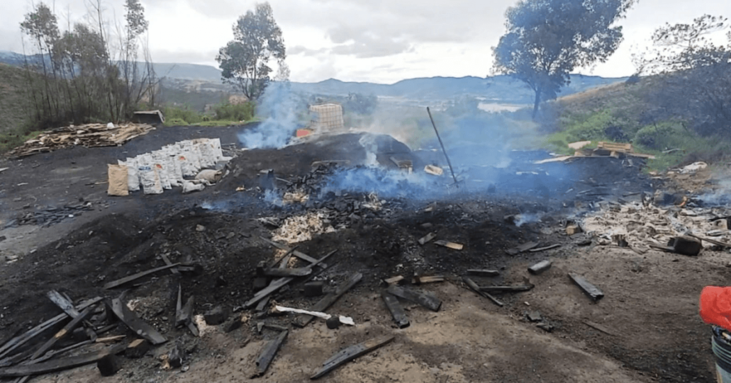 Carbón vegetal, cenizas y residuos de madera humeantes en un área intervenida al aire libre, con sacos apilados y vegetación alrededor en una zona rural.
